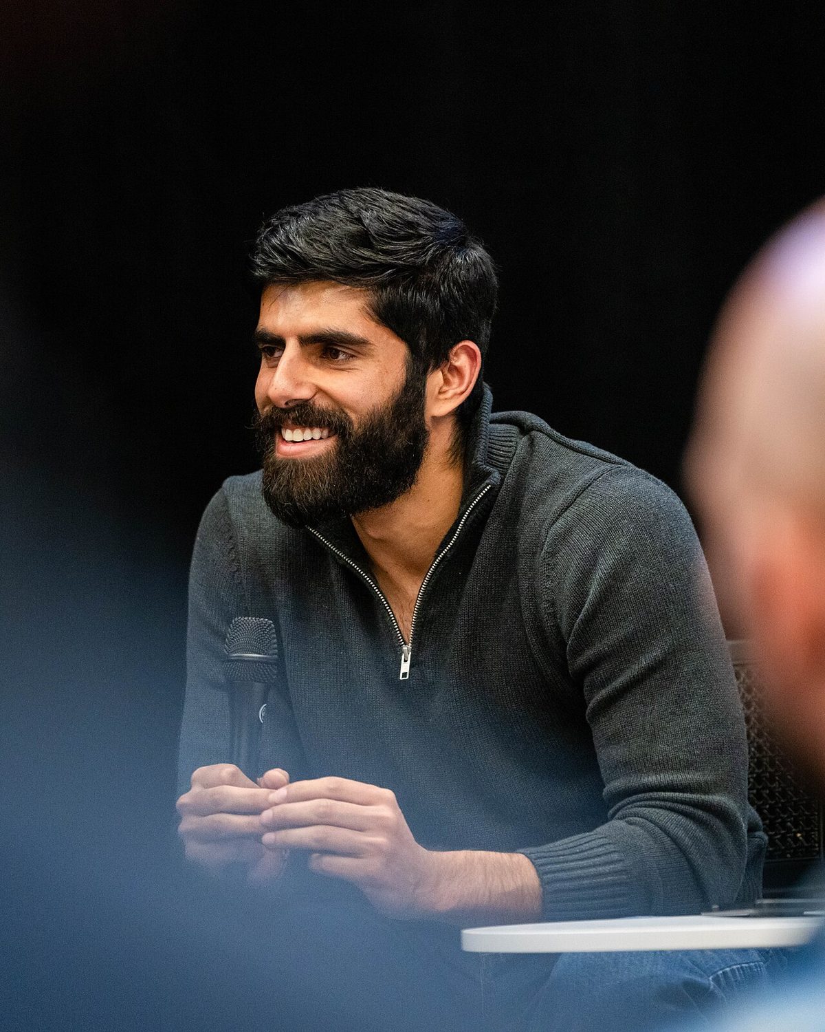 A bearded man in a dark quarter-zip sweater, smiling and looking off to one side, hands together around a microphone at a low table