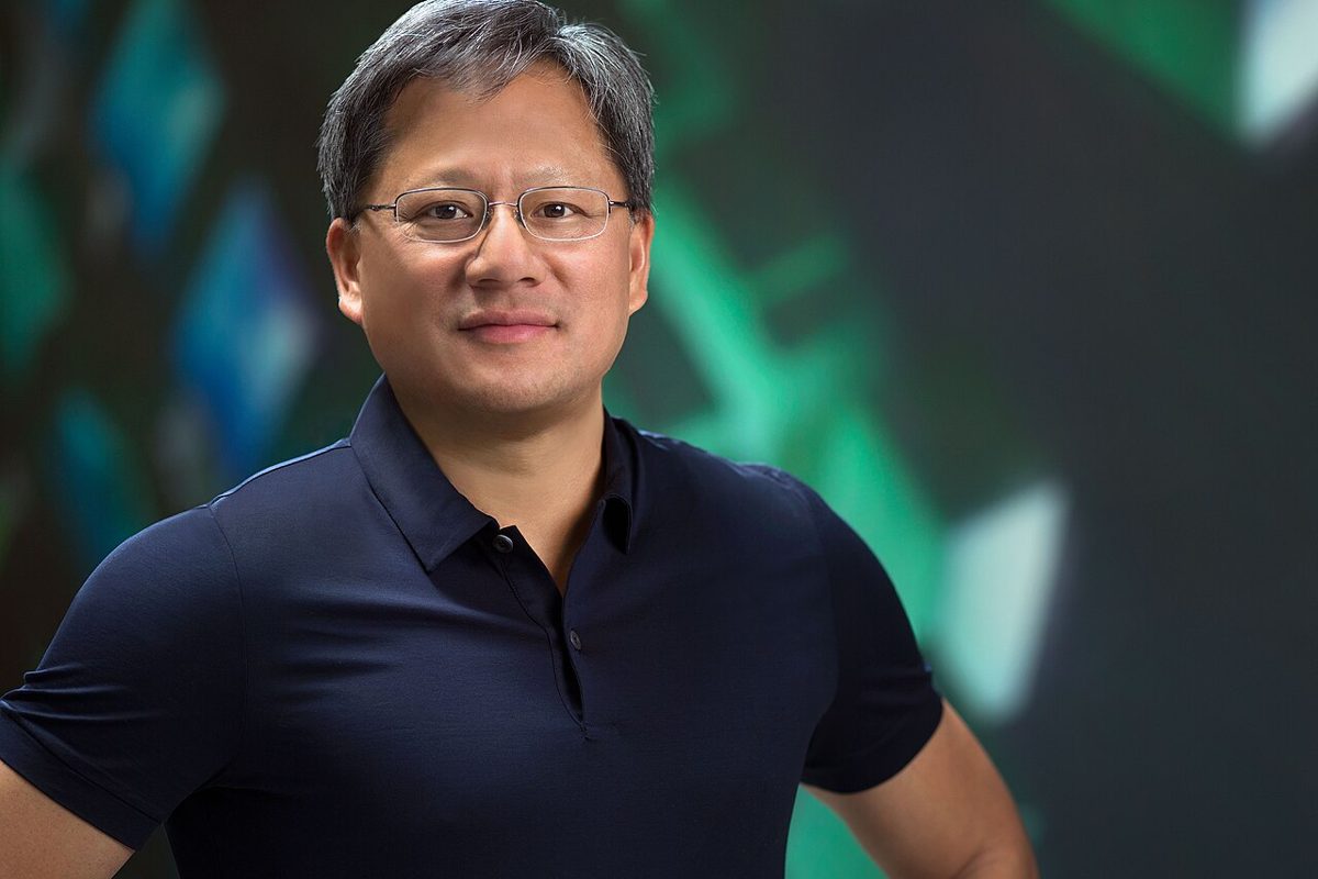 A man with gray hair and rimless glasses, in a dark navy polo shirt, standing with his hands on his hips in front of a blurred green and blue backdrop