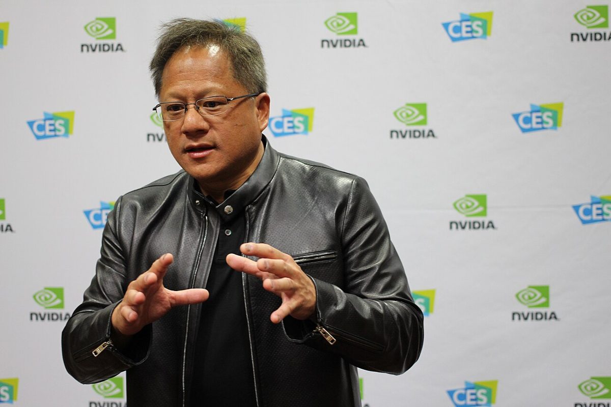 Jensen Huang in glasses and a black leather jacket, gesturing with both hands as he talks, in front of a white backdrop printed with Nvidia and CES logos