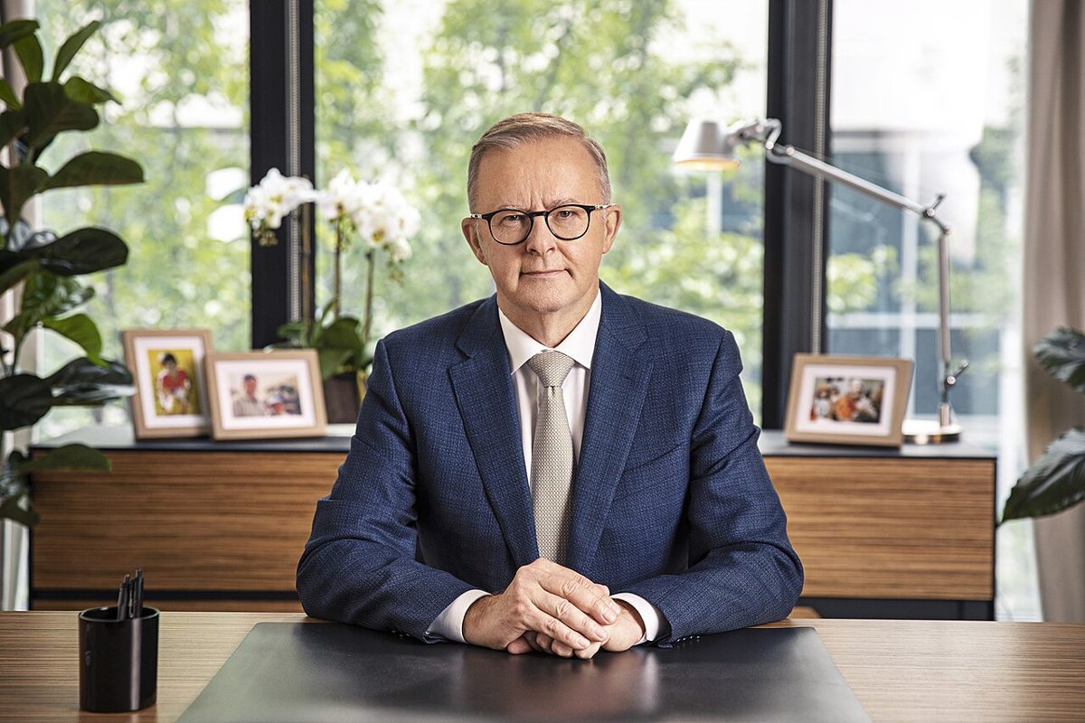 Anthony Albanese in glasses and a blue suit, seated at a desk with his hands folded, framed photos and a desk lamp behind him