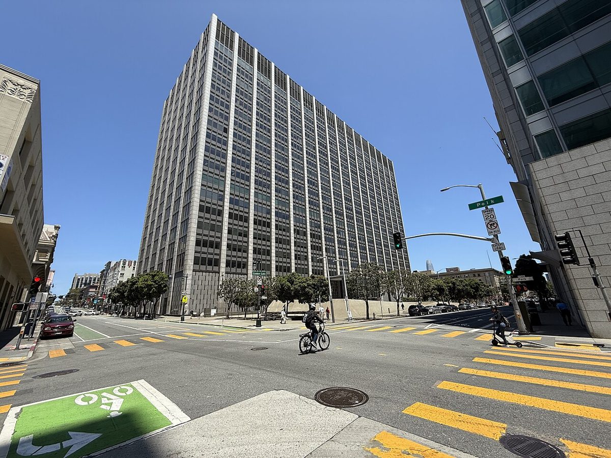 A tall gray office tower with long vertical window bands stands on a sunny street corner, with a cyclist crossing the intersection in front of it