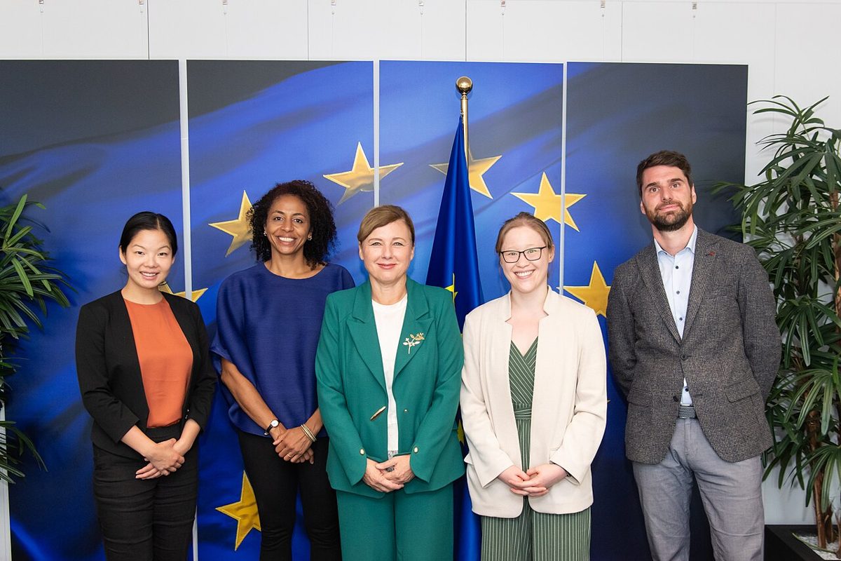 Five people standing in a row and smiling for a group photograph in front of a European Union flag and a wide backdrop of gold stars on deep blue
