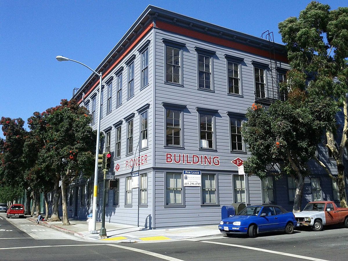 A three-story grey clapboard building on a street corner with PIONEER BUILDING painted in red letters, For Lease signs in the windows and flowering trees along the sidewalk