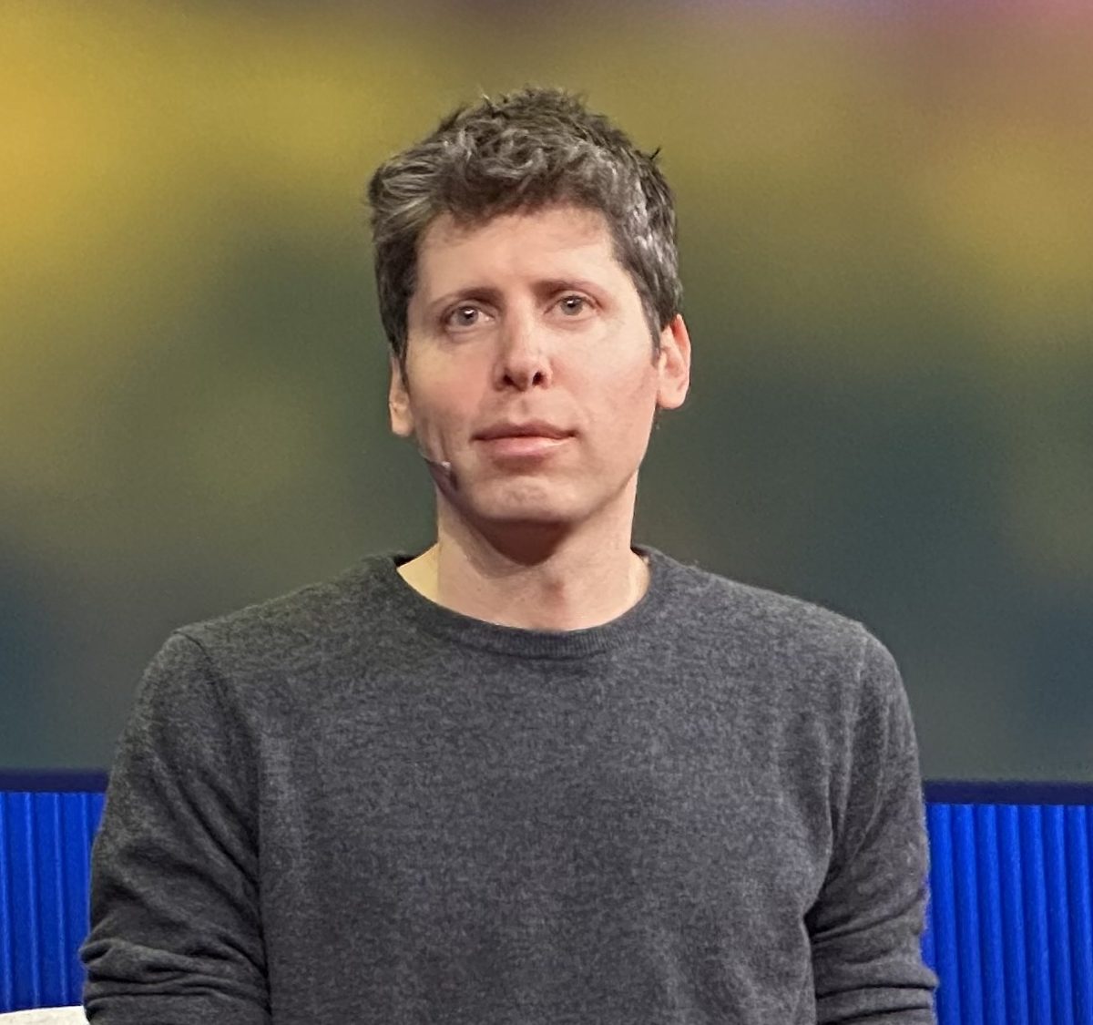A man in a gray crew-neck sweater with a thin headset microphone across his cheek, looking off camera against a blurred yellow and green backdrop
