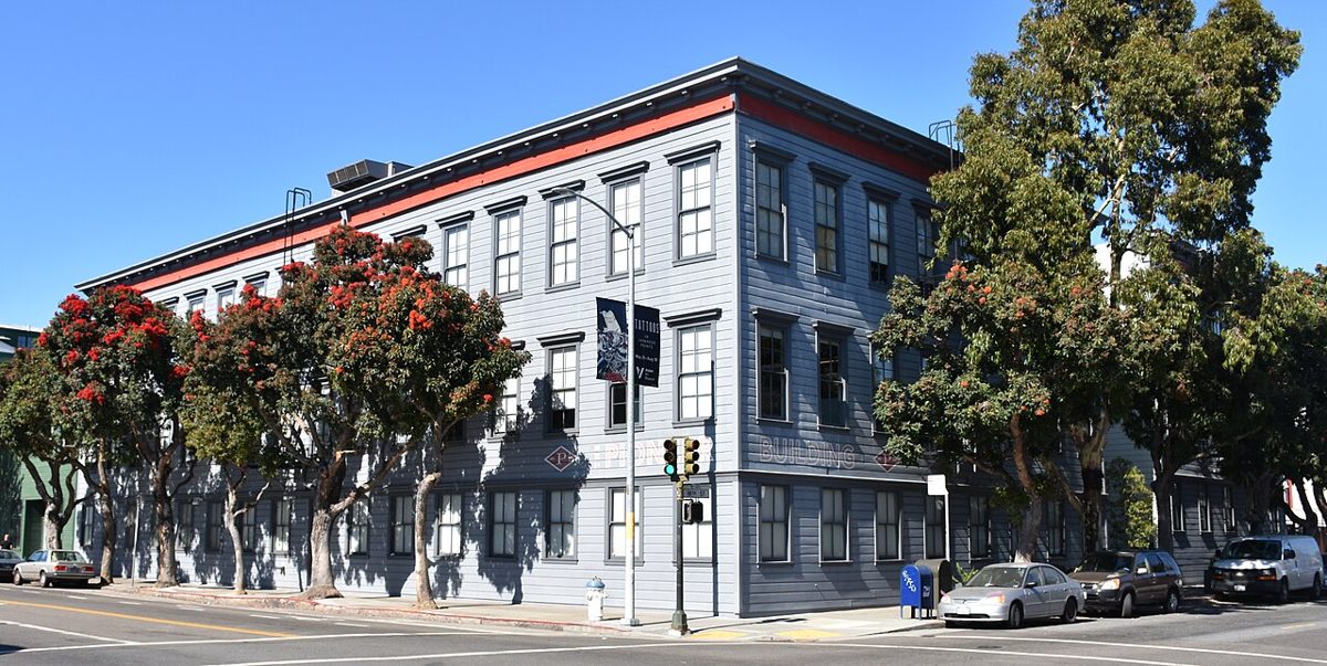 A three-story grey clapboard building on a sunny street corner, with red-flowering trees along the pavement and cars parked at the kerb