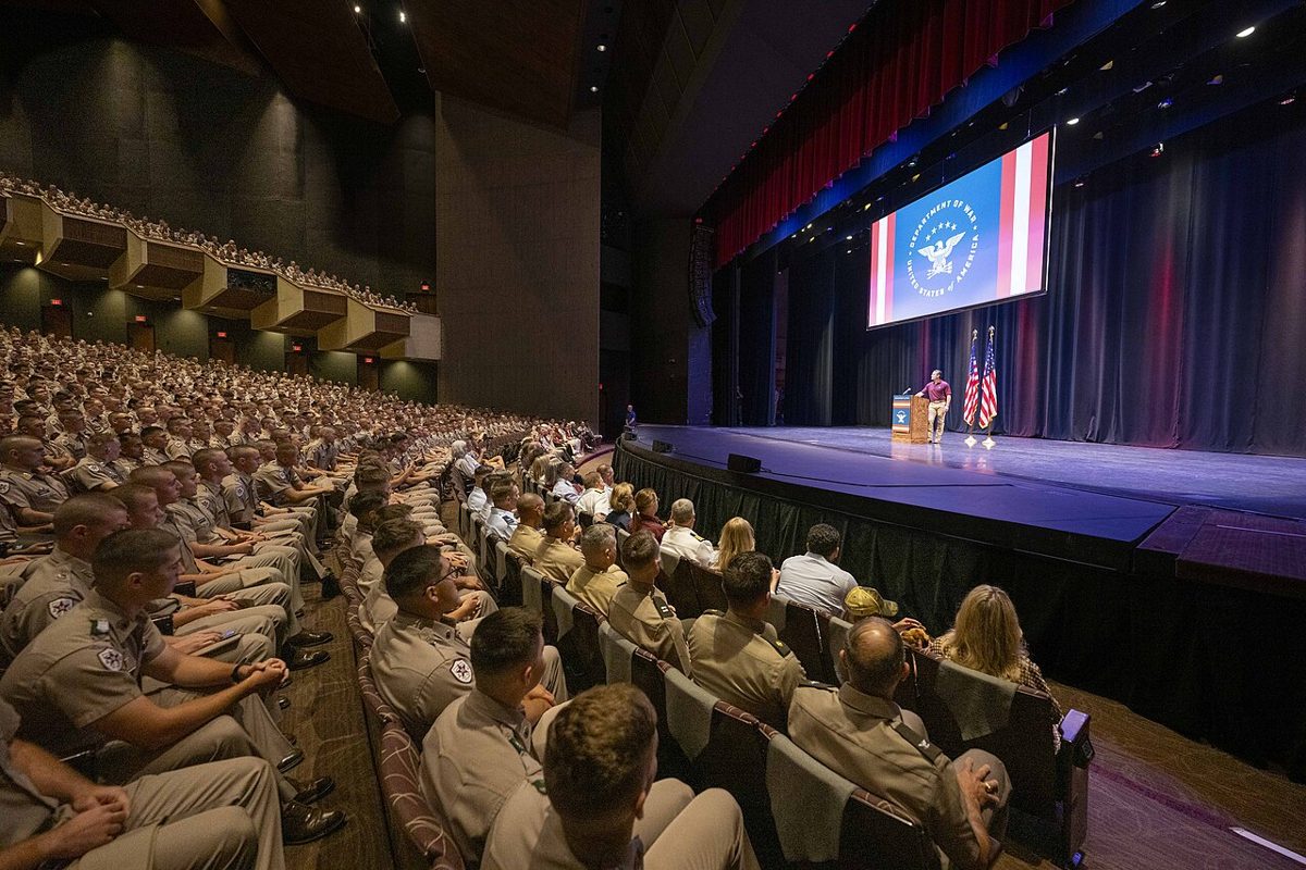 Pete Hegseth at a lectern on a large stage under a Department of War seal, facing a hall full of cadets in khaki uniforms