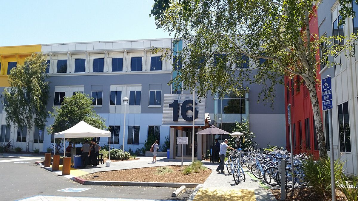 A three-story office building with a large number 16 over its entrance and rows of blue bicycles parked under a tree