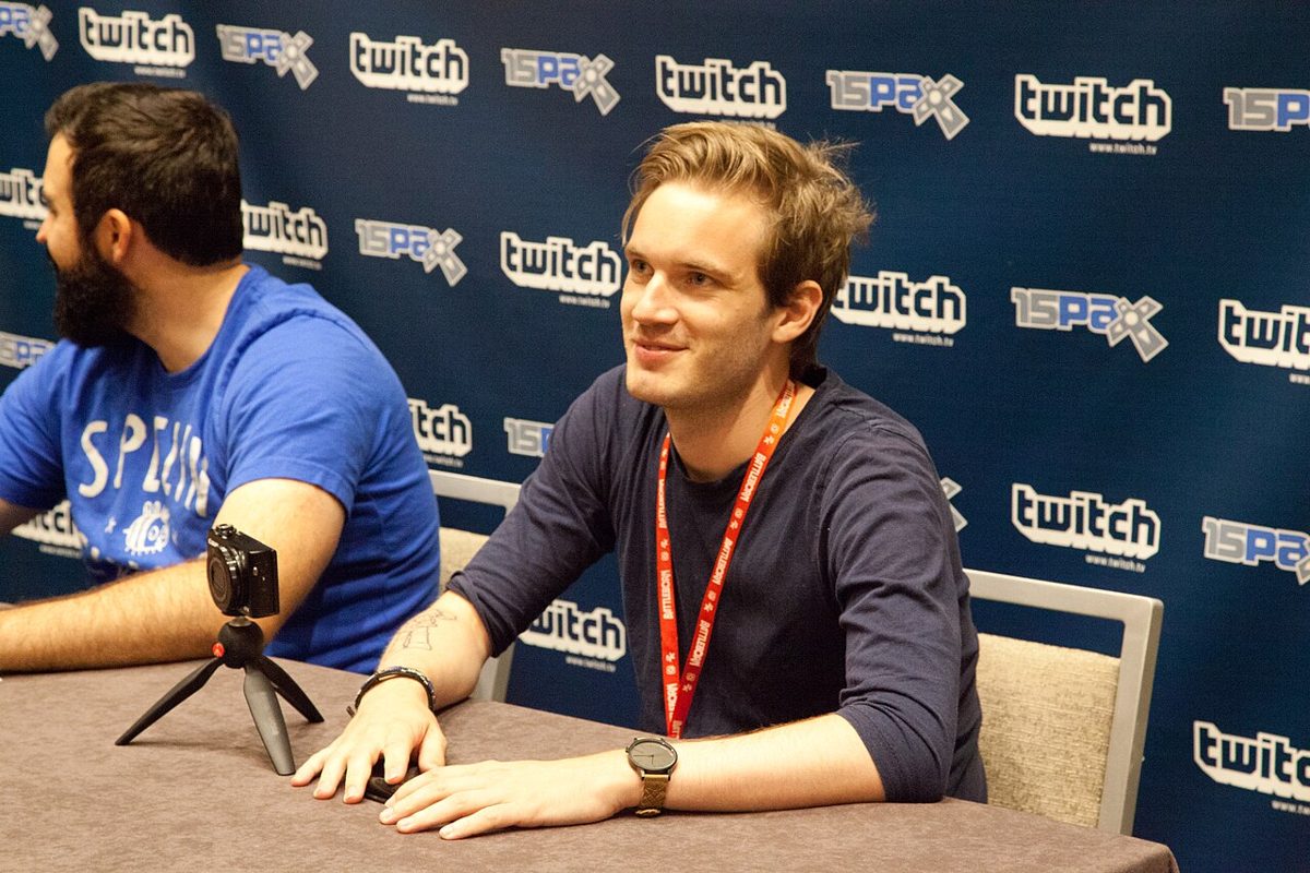 A young man with swept-back blond hair and a red lanyard sits smiling at a table in front of a Twitch and PAX backdrop