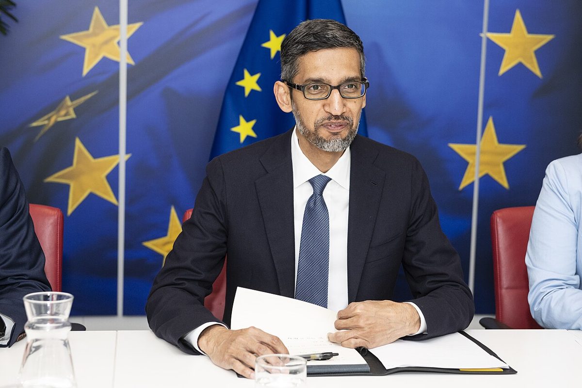 A bearded man in glasses, a dark suit and blue tie, seated at a white table with papers, in front of a blue EU flag backdrop