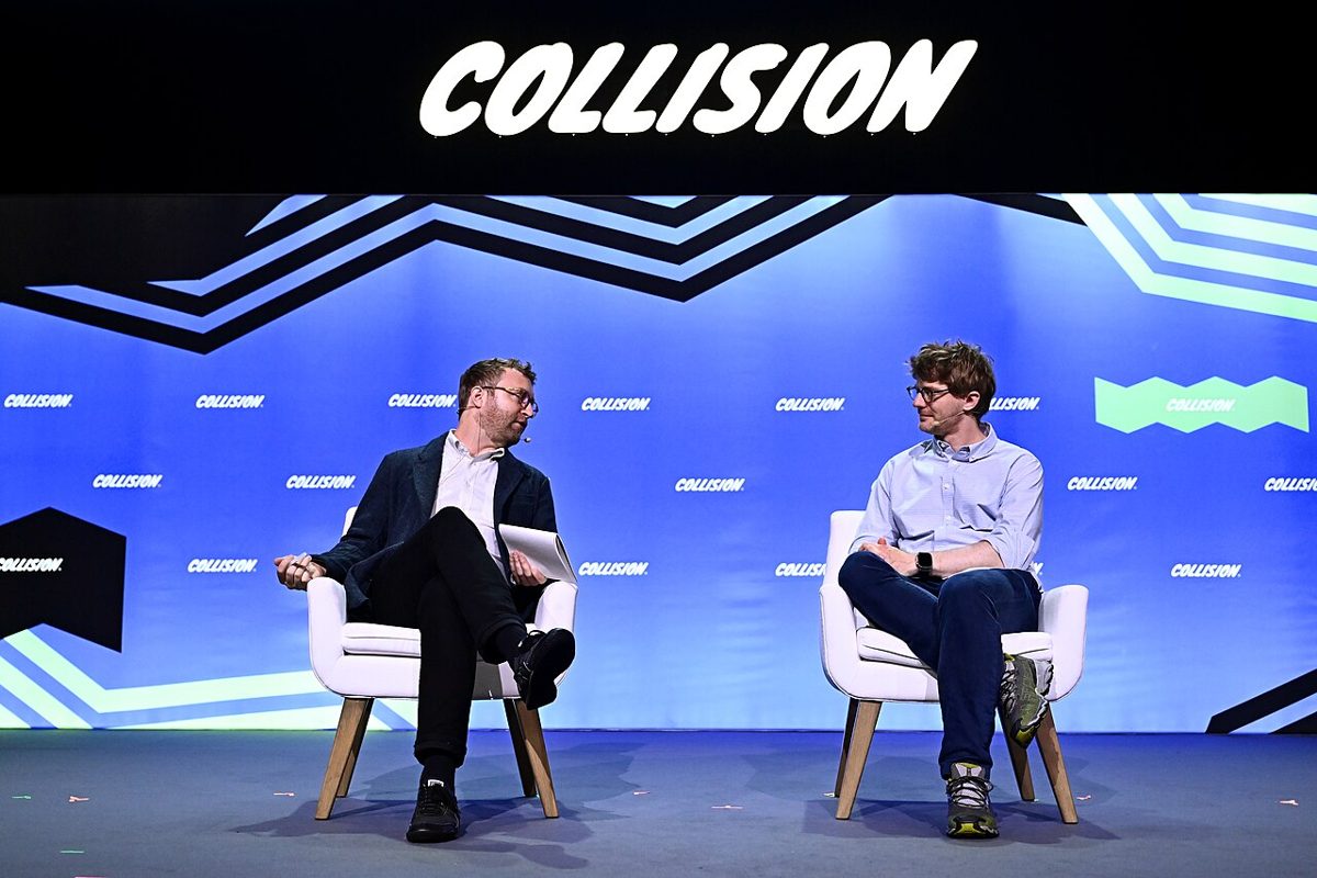 Two men talking in white armchairs on a blue stage under a large Collision sign