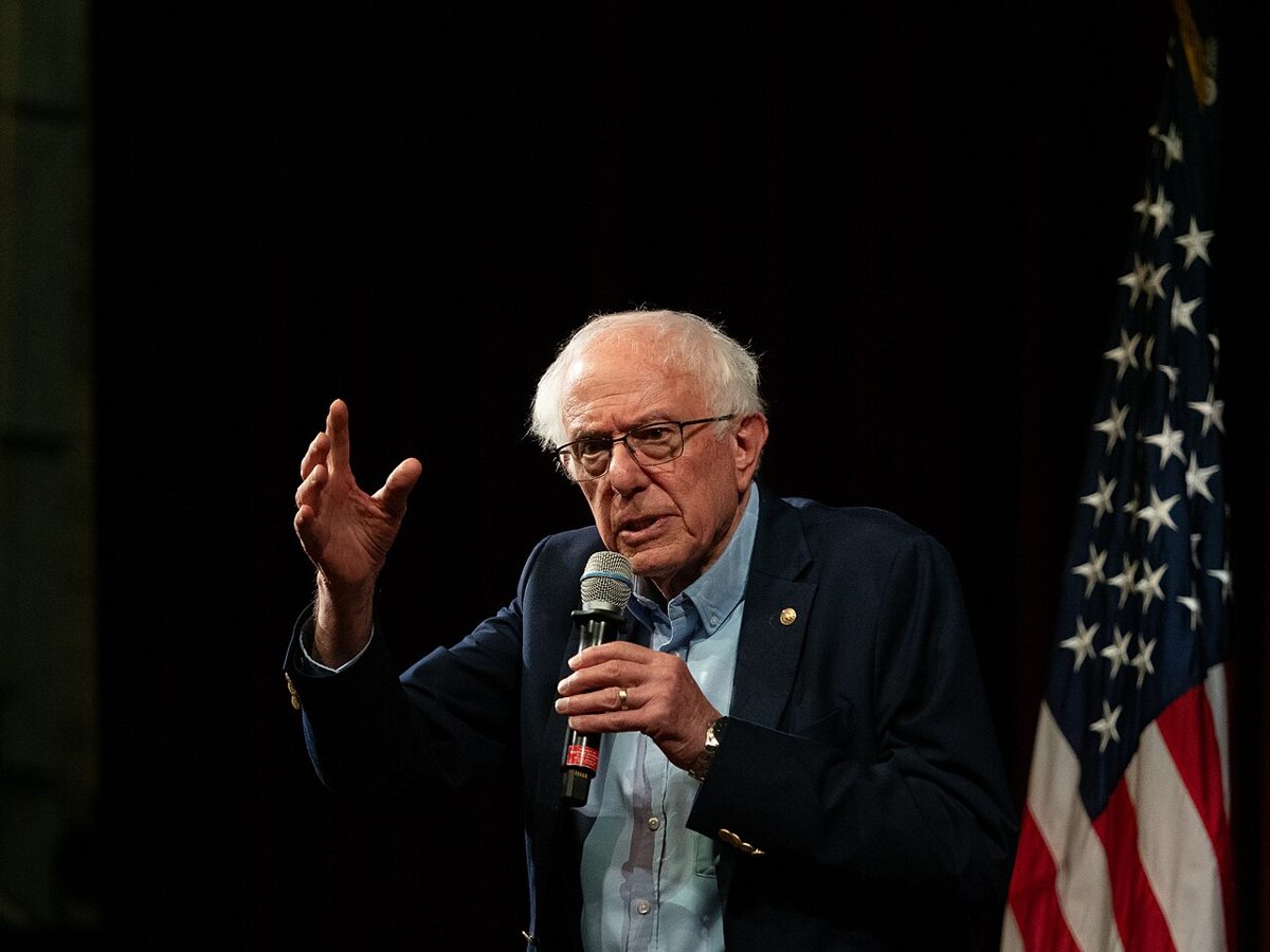 Bernie Sanders in glasses and a navy blazer speaks into a microphone and gestures with his left hand, an American flag behind him on a dark stage