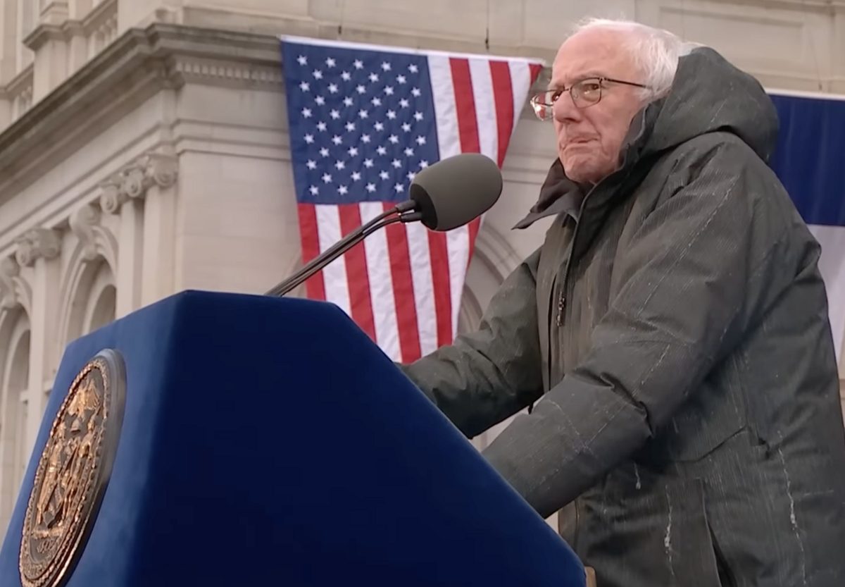 An elderly man with white hair and glasses, in a dark winter coat, speaks at a blue lectern with a gold seal in front of an American flag