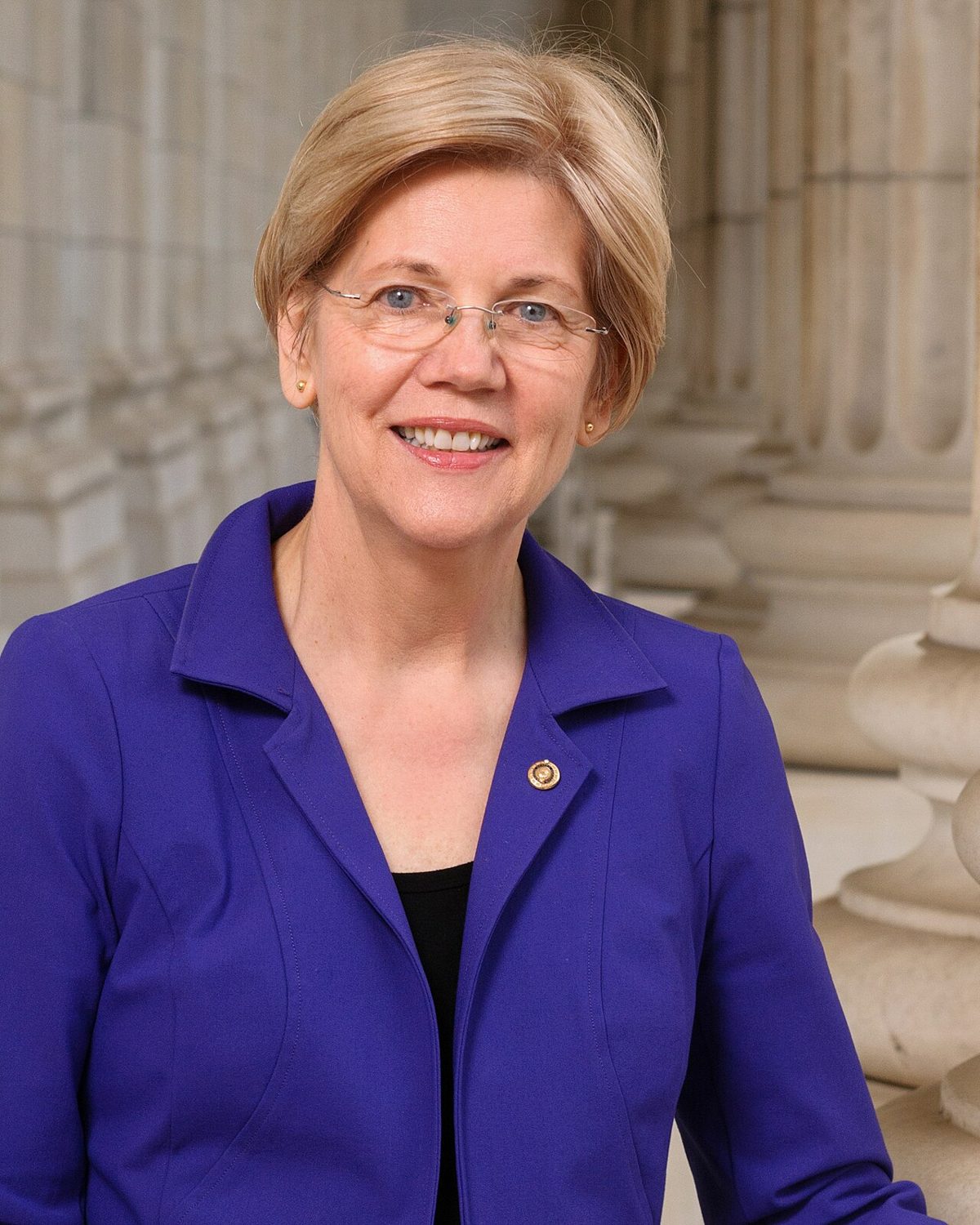 Elizabeth Warren, smiling, in rimless glasses and a purple jacket with a Senate pin, standing in front of a row of stone columns