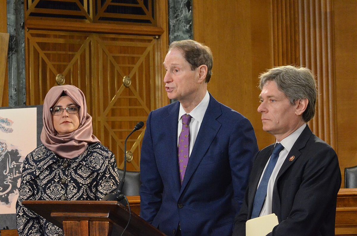 A gray-haired man in a navy suit and purple tie speaks beside a podium in a wood-paneled room, between a woman and another man