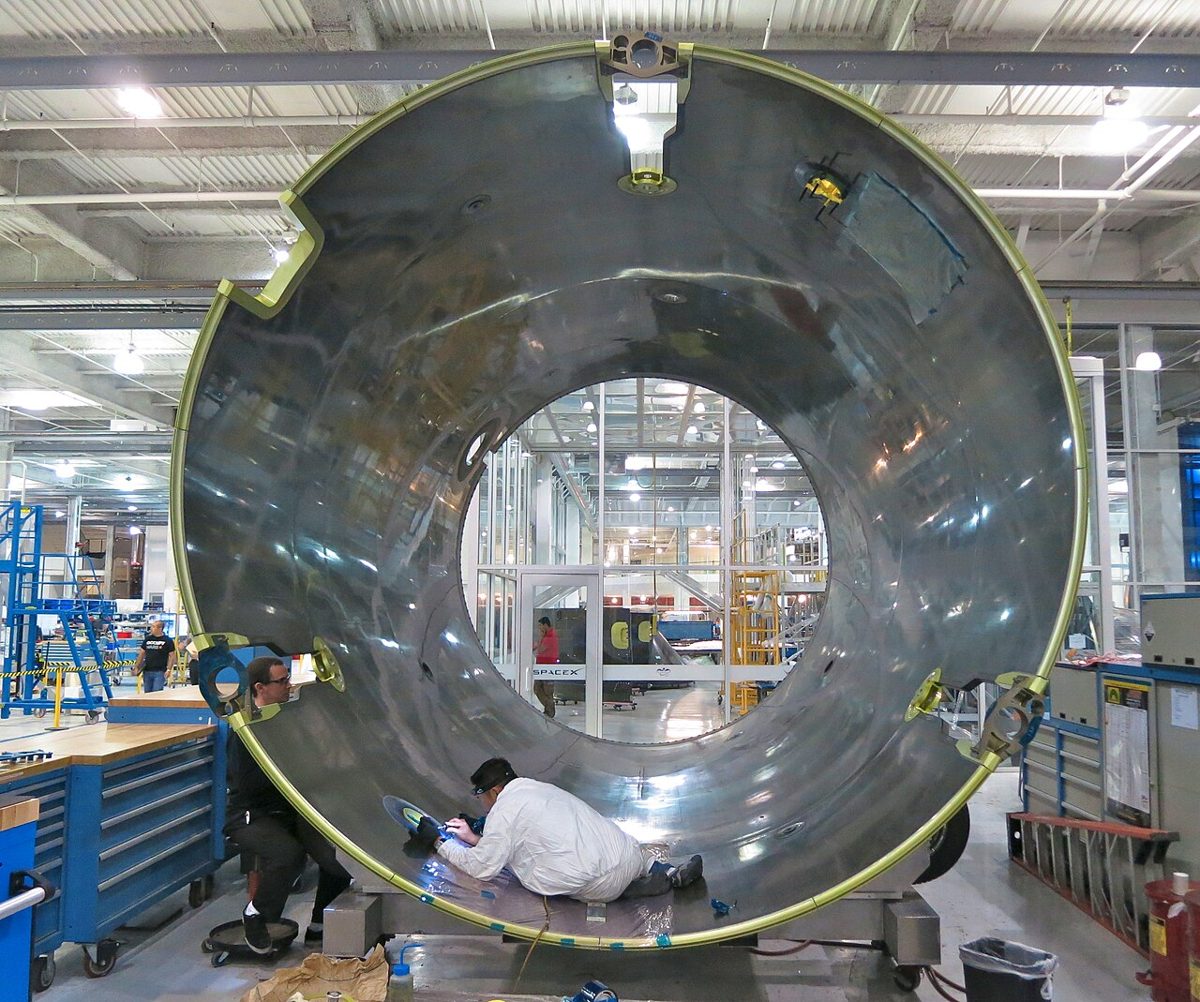 A worker kneels inside a large polished metal rocket section on a factory floor while another sits beside it, with blue tool cabinets and glass-walled rooms behind