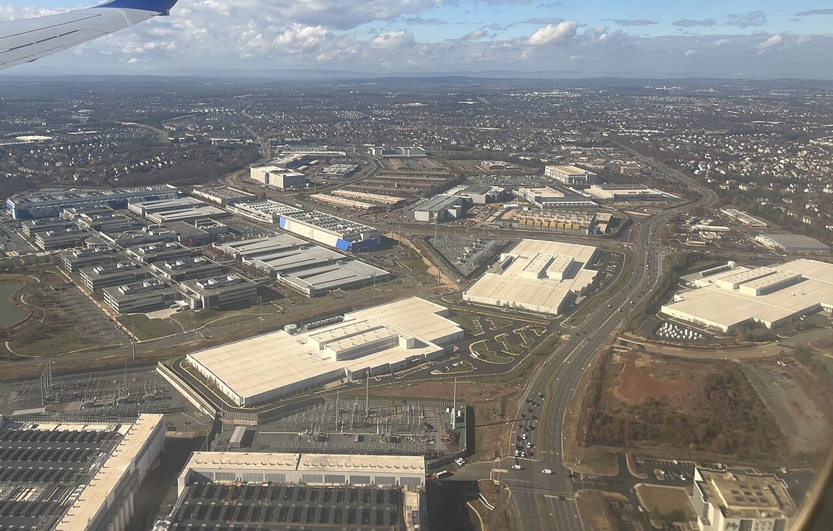 An aerial view of dozens of flat white warehouse-sized buildings with rooftop cooling units, laid out beside electrical substations and a highway, with suburbs beyond