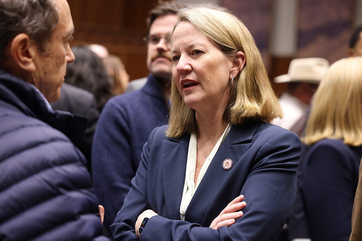 A woman with shoulder-length blonde hair and a navy blazer, arms folded, talking with a man in a padded jacket in a crowded wood-paneled room