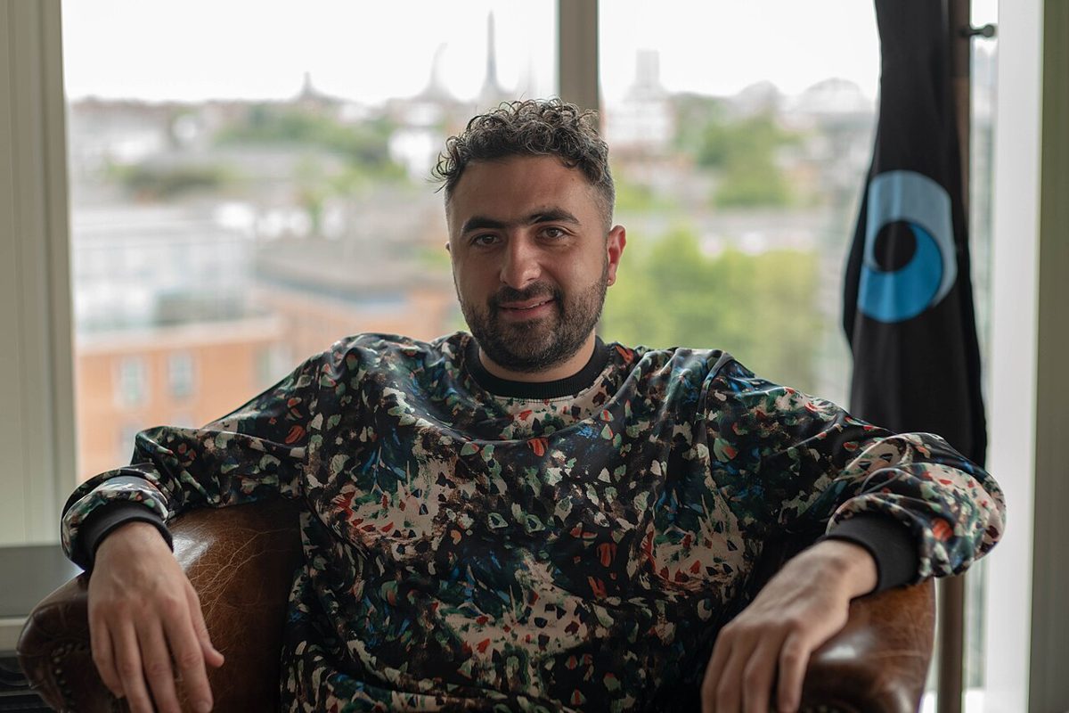 A bearded man in a patterned sweatshirt sits in a leather armchair beside a window looking out over a low city skyline