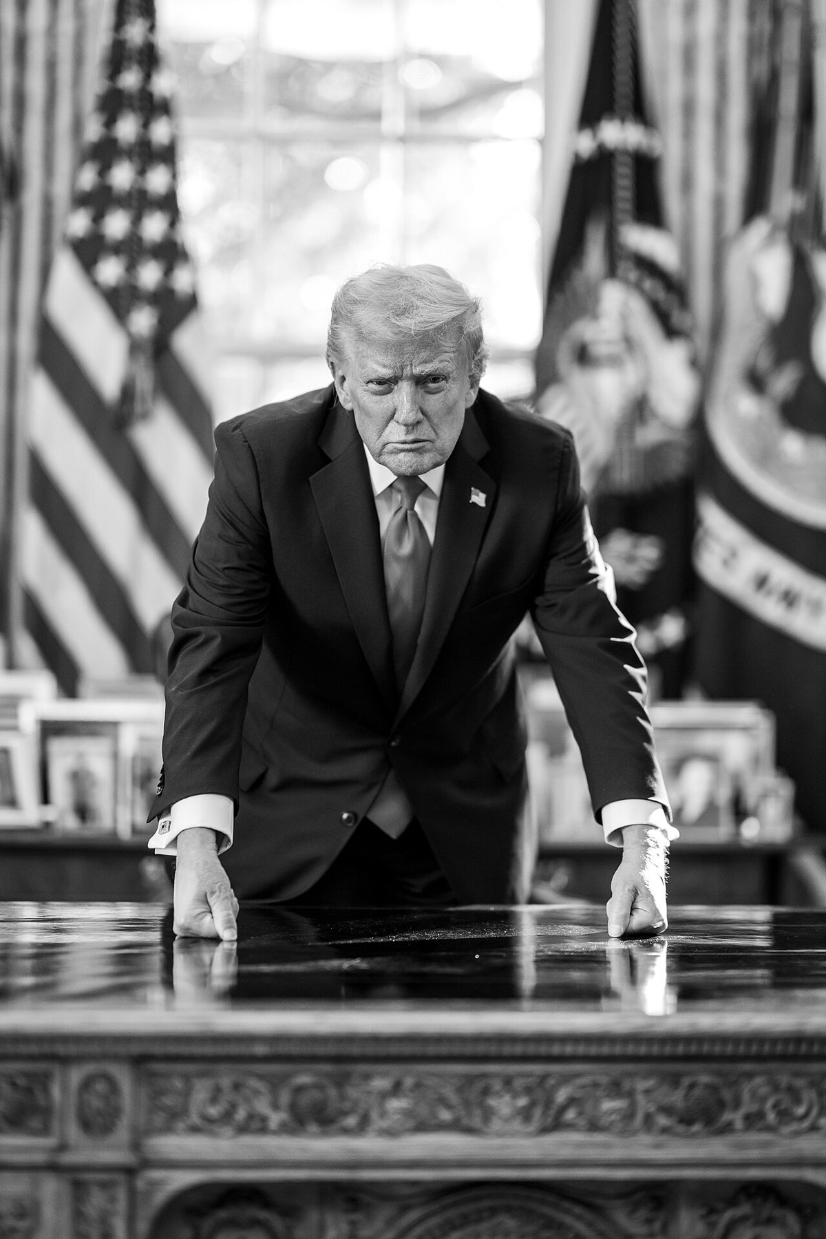 Donald Trump in a dark suit leans forward with both hands flat on a polished desk, flags and a bright window behind him, in black and white