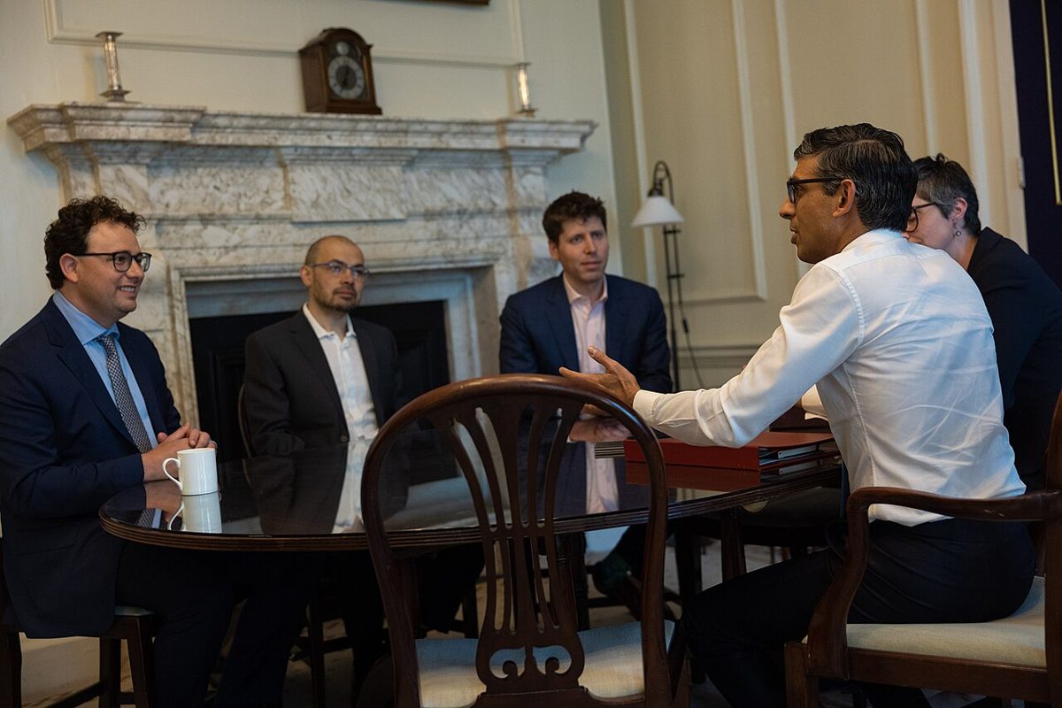 Five people at a glass-topped table by a marble fireplace, a smiling man in glasses on the left