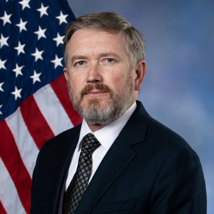 Thomas Massie, gray-haired with a gray beard, in a dark suit and patterned tie, in front of an American flag and a blue backdrop