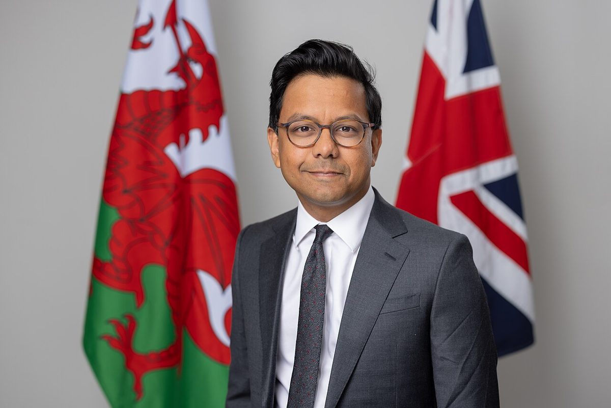 A man with short dark hair and round glasses, in a gray suit, white shirt and dark tie, standing in front of a Welsh flag and a Union flag