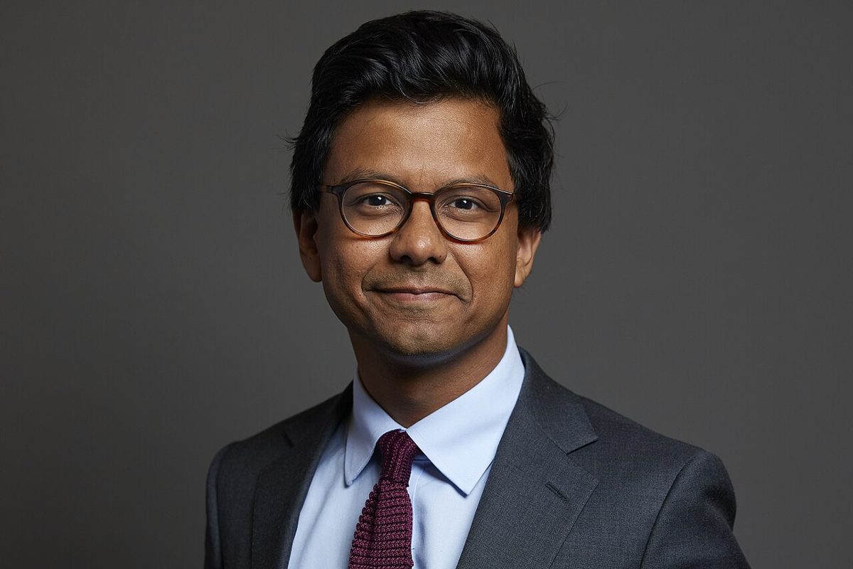 A man with dark hair and round tortoiseshell glasses, in a gray suit, light blue shirt and knitted burgundy tie, smiling slightly in front of a plain gray backdrop