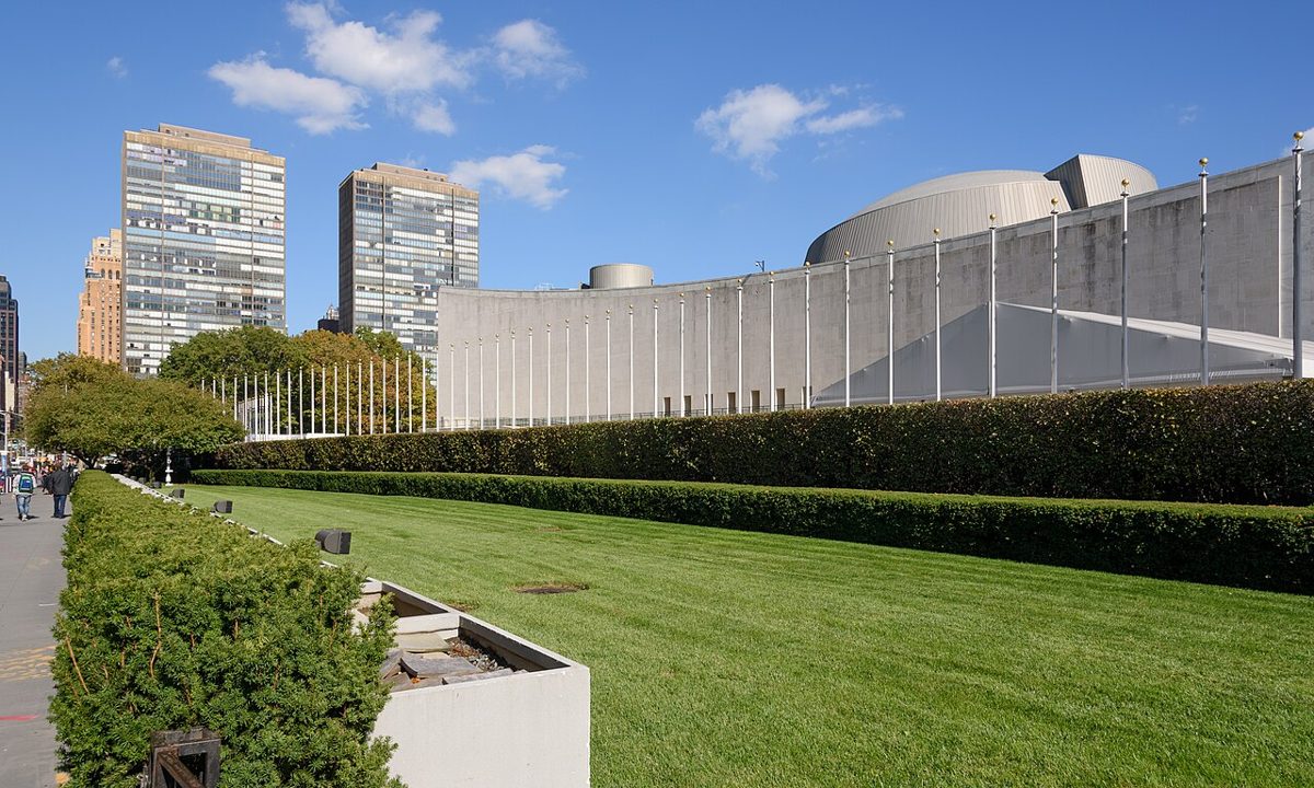 A row of bare white flagpoles behind a clipped hedge and a wide lawn, with the low concrete General Assembly Building and its shallow dome beyond