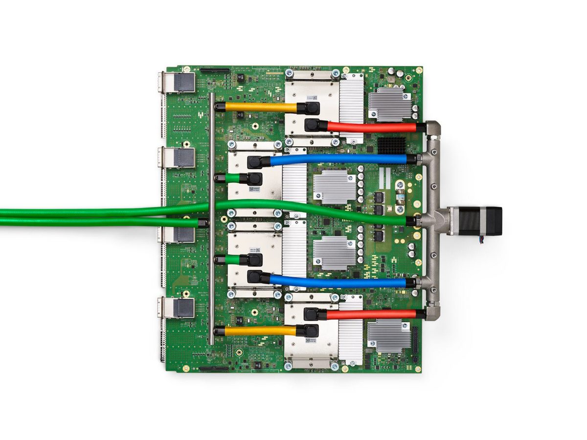 A green circuit board seen from above, with four chip packages under metal heat spreaders and red, blue, yellow and green coolant hoses running to a manifold on one side