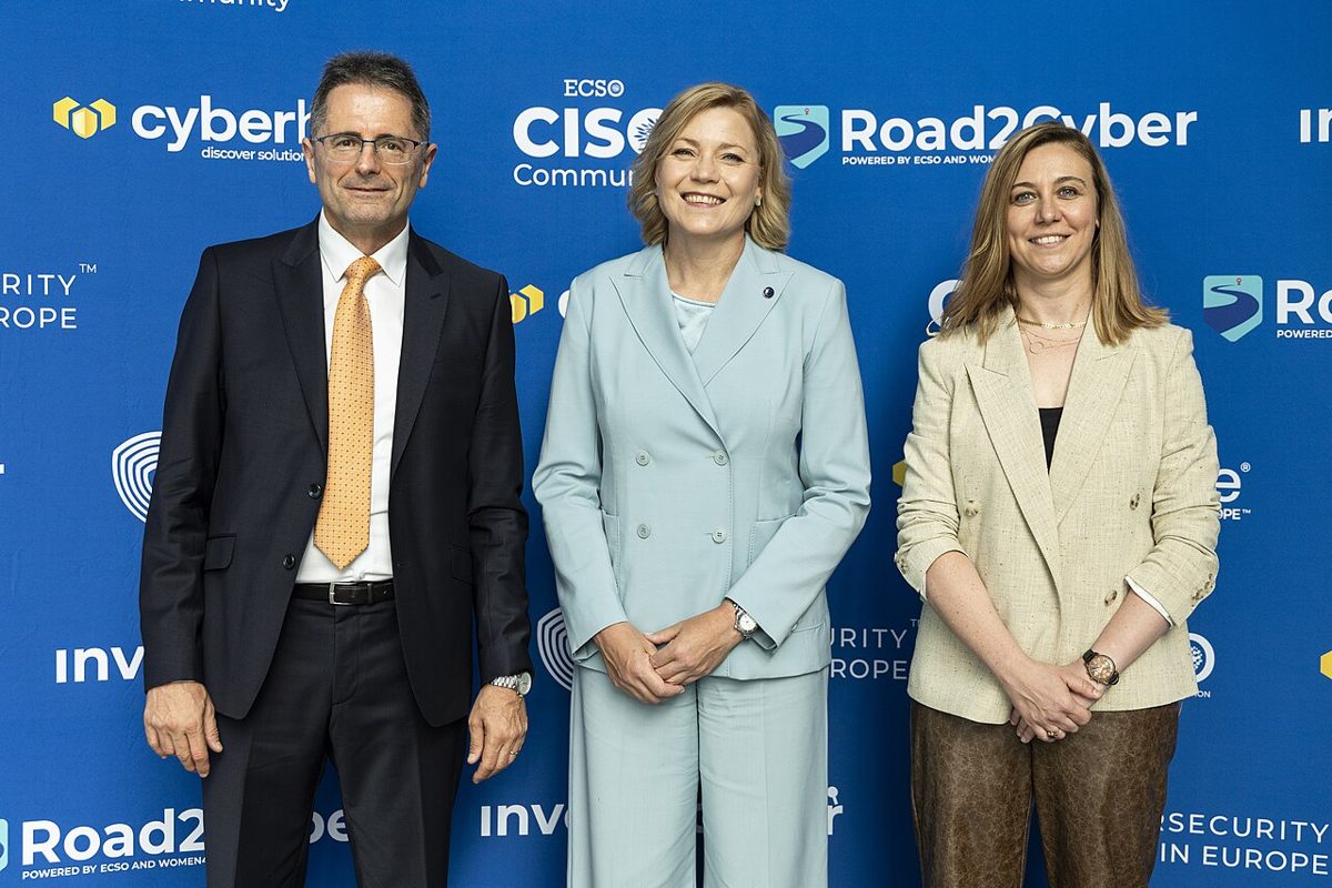 A smiling blonde woman in a pale blue suit stands between a man and a woman in front of a blue event backdrop