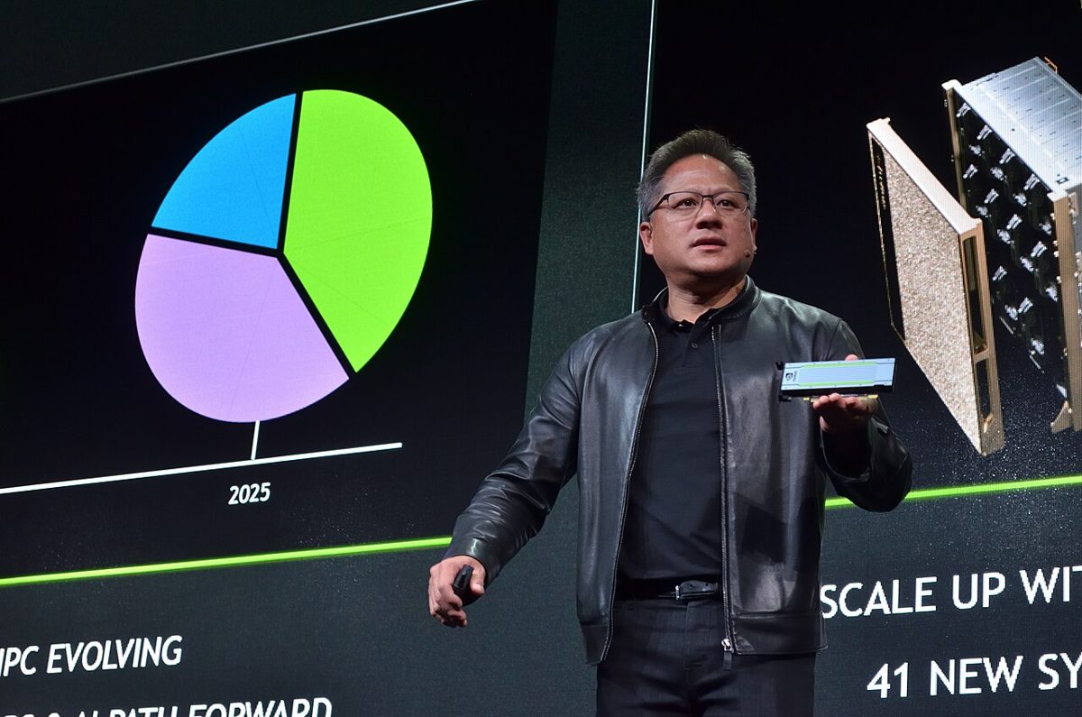 Jensen Huang in a black leather jacket and glasses on stage, holding up a small circuit board in front of a large screen showing a pie chart