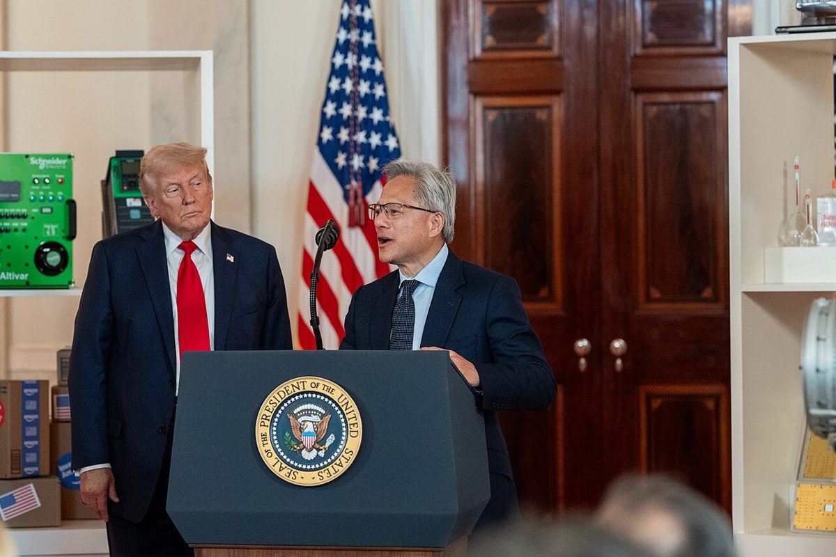 Jensen Huang, in glasses and a dark suit, speaks at a lectern with the presidential seal while Donald Trump, in a red tie, stands behind him listening