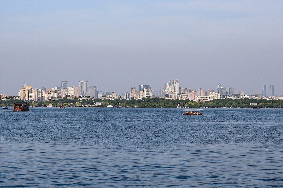 A wide lake with small wooden tour boats on the water and a long line of high-rise buildings along the far shore under a hazy blue sky.