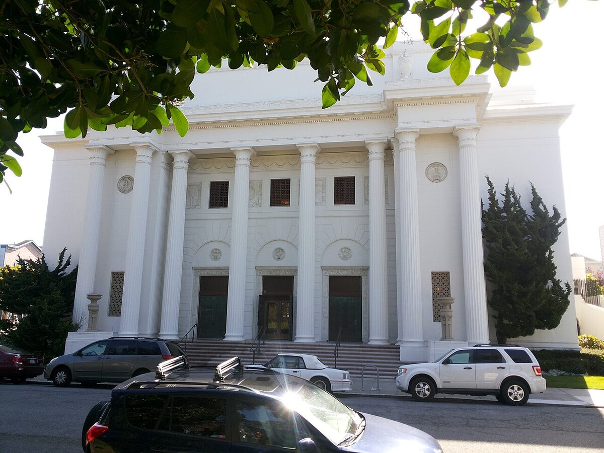 A white neoclassical building with tall fluted columns and three doorways at the top of wide steps, with cars parked along the street in front and leaves overhanging the top of the frame.