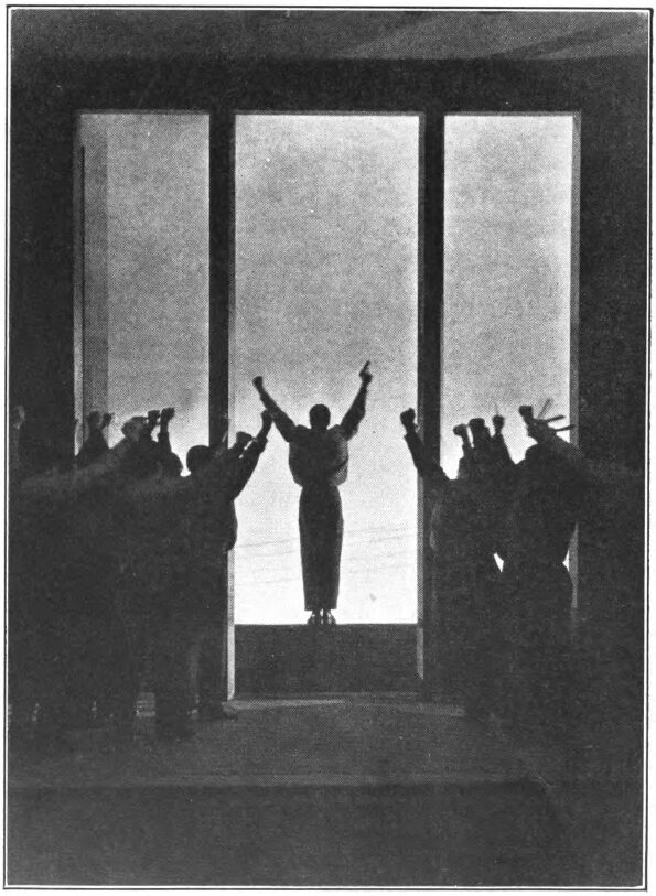 Stage photograph of silhouetted figures raising clenched fists toward one figure who stands with both arms up against tall, bright windows.