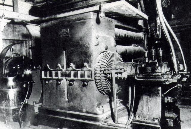 Old black-and-white photograph of a massive iron dynamo with a large ribbed armature, gauges and pipes, in a cramped engine room.