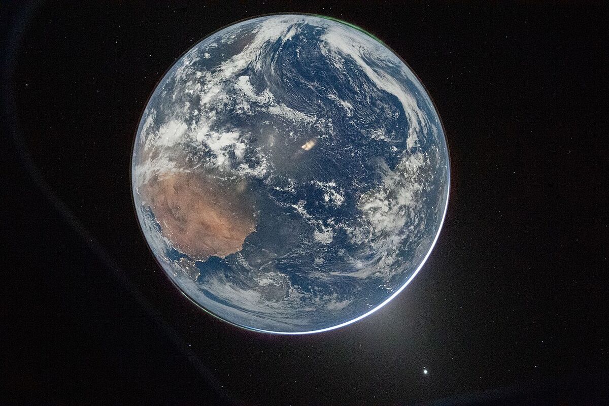 The whole Earth seen through a spacecraft window: blue ocean and white clouds, with the red-brown land of Australia at lower left and a thin bright edge of light.