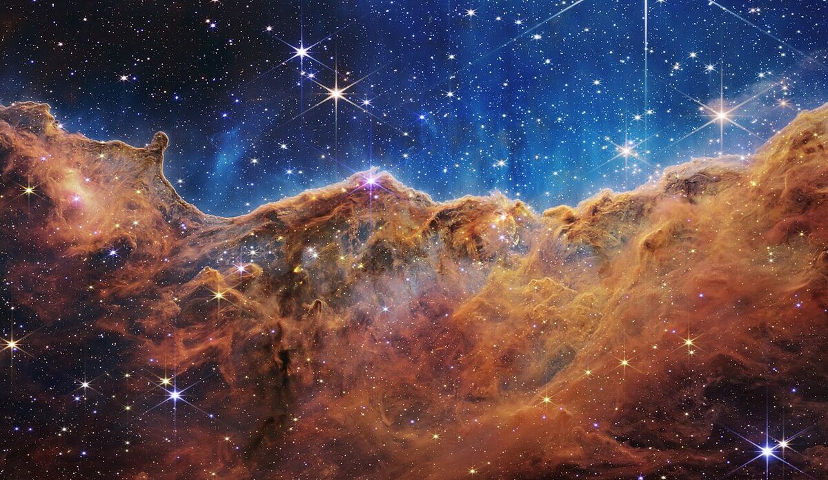 Glowing orange-brown clouds of gas and dust rising like a jagged mountain ridge beneath a deep blue sky full of bright, spiky stars.