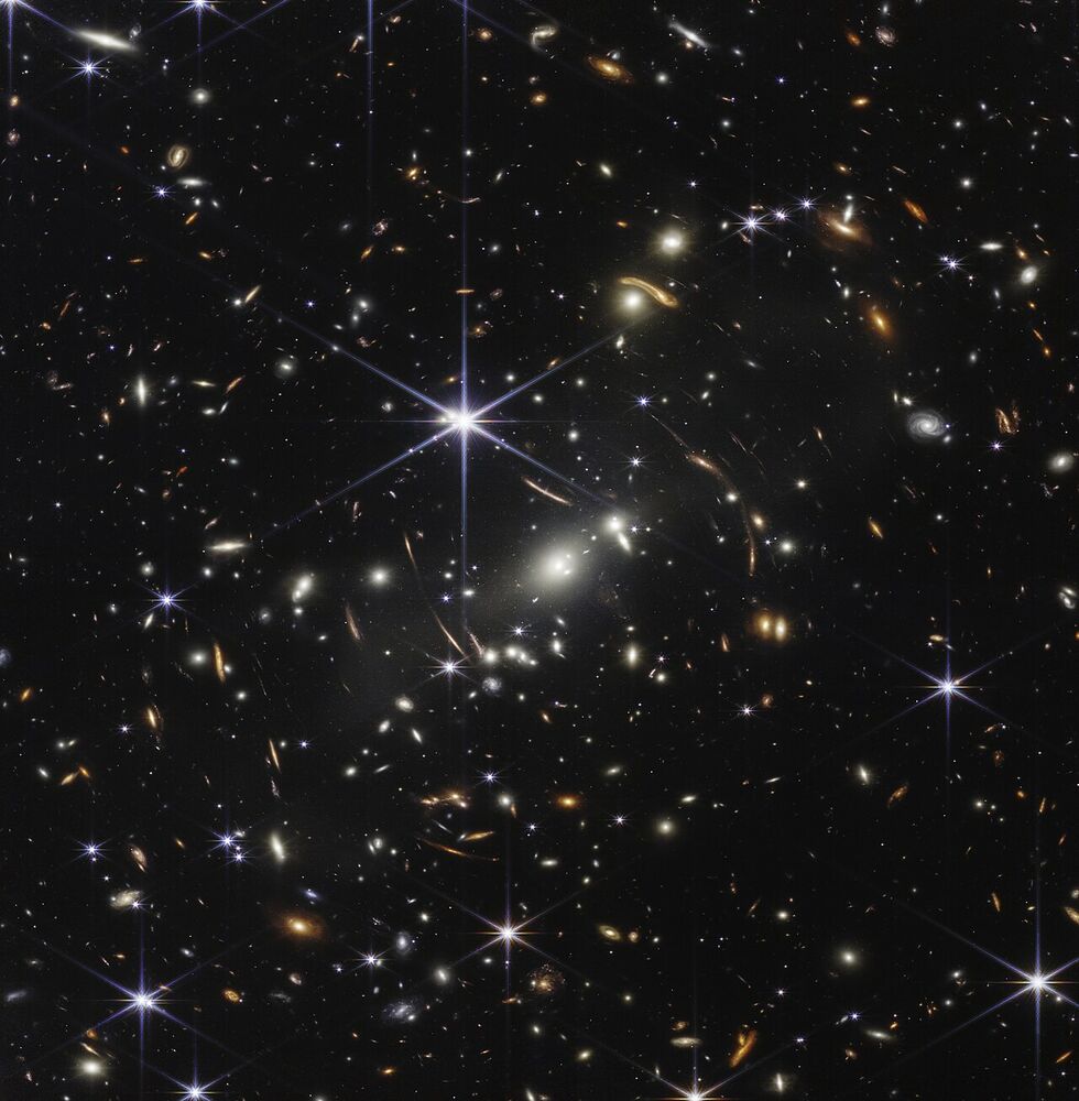Thousands of tiny galaxies scattered across black space, some stretched into thin orange arcs, with a few bright stars showing six-pointed spikes.