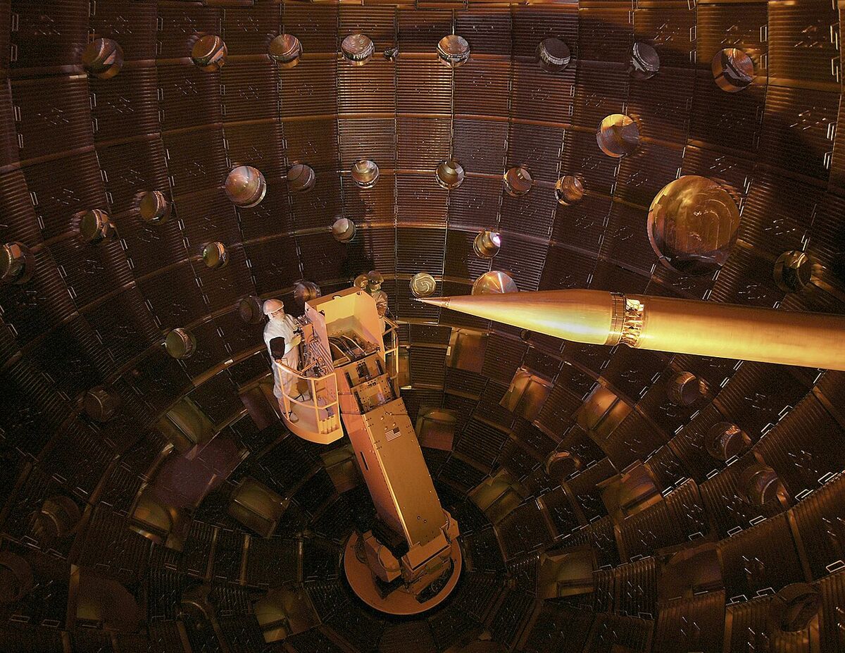 Inside a huge copper-colored spherical chamber lined with round ports, a technician on a lift works near a long pointed arm aimed at the center.