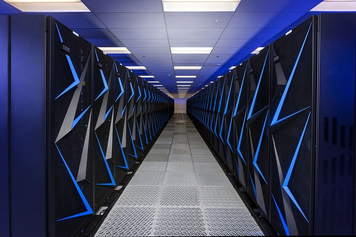 A long aisle between two rows of black supercomputer cabinets marked with bright blue angular stripes, under a blue-lit ceiling.