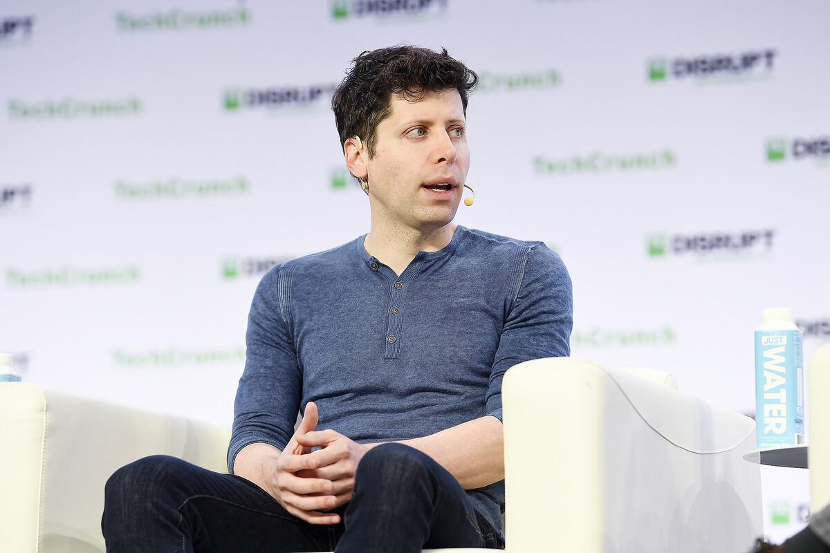 Sam Altman in a blue henley and jeans, wearing a headset microphone, sitting in a white armchair on a stage in front of a TechCrunch Disrupt backdrop