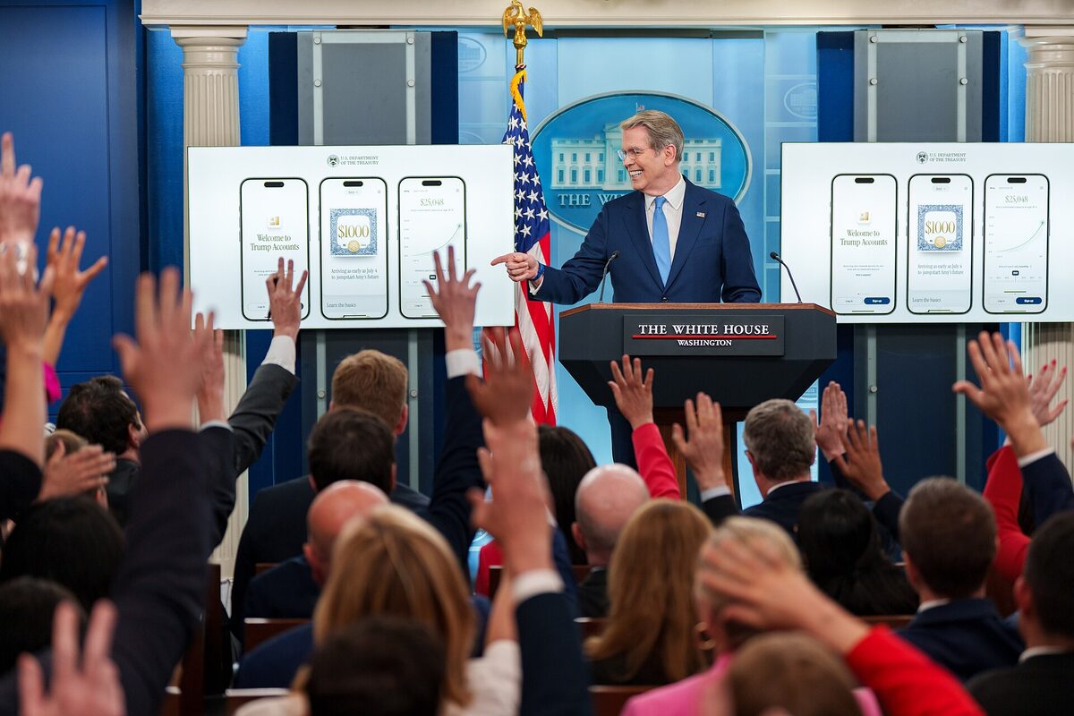 A smiling man in glasses and a blue suit points from a White House lectern toward reporters raising their hands, with screens of phone app mockups on either side of him