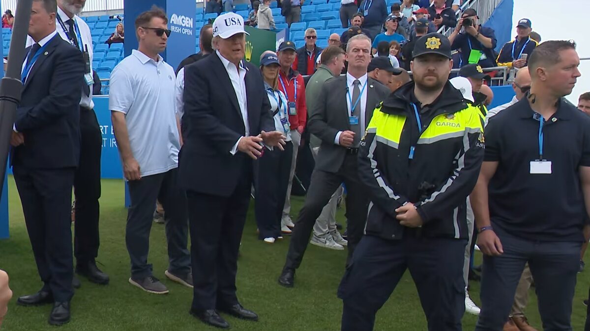 Donald Trump in a white USA cap and dark suit standing on a golf course lawn beside security staff and an Irish police officer in a yellow Garda jacket, with spectators in a blue grandstand behind