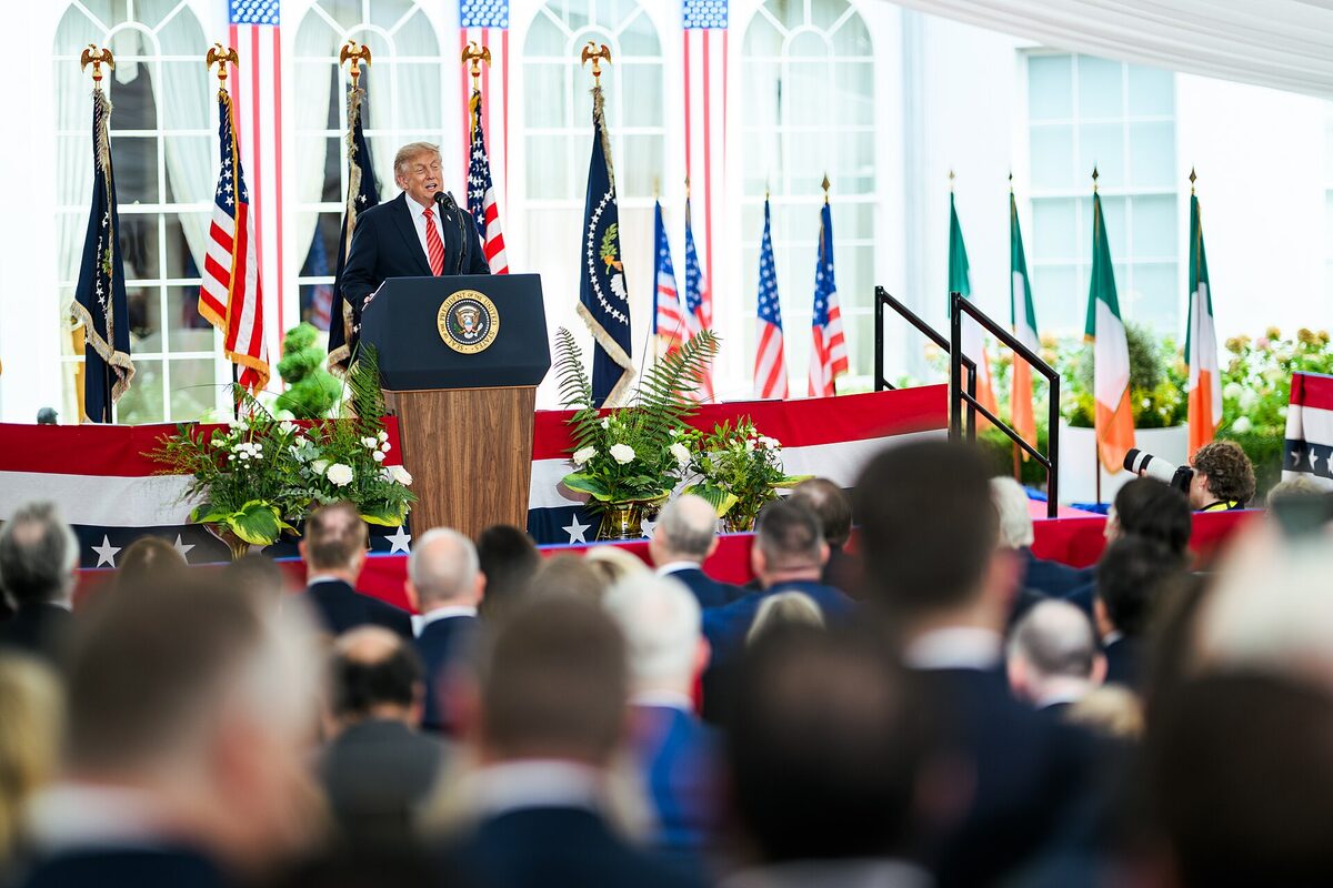 Donald Trump speaking at a lectern bearing the presidential seal under a white tent lined with American and Irish flags, with rows of seated guests blurred in the foreground