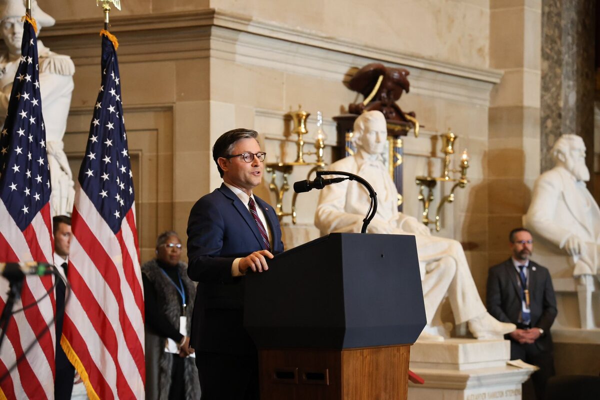 Mike Johnson in glasses and a navy suit speaking at a lectern between American flags, with white marble statues in a stone hall behind him